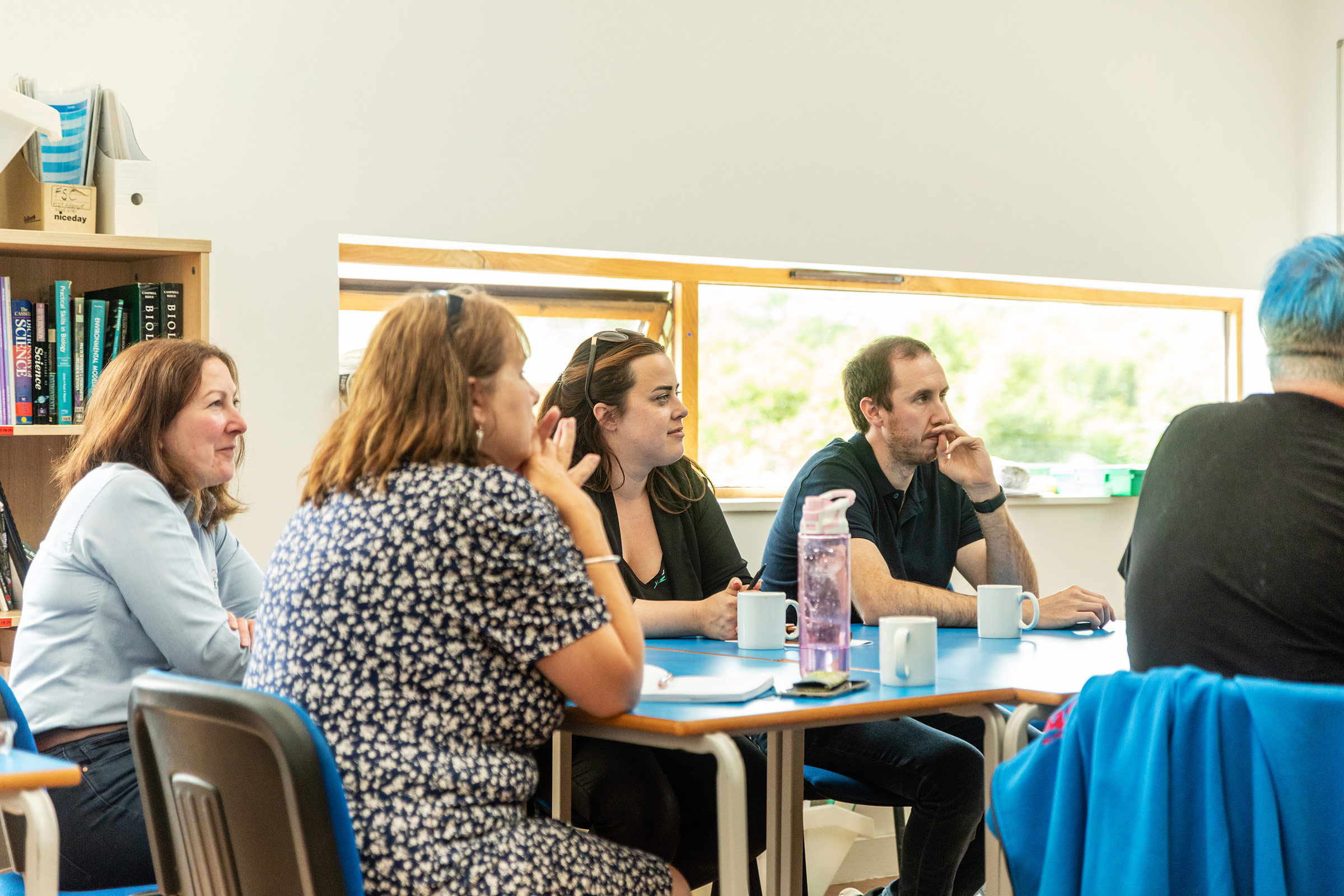 Group of people round a table looking at a speaker. 3 women, two men. All white, looking interested in subject.