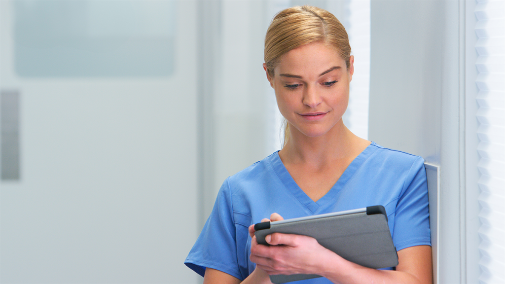 Woman in scrubs leaning on wall working on a tablet