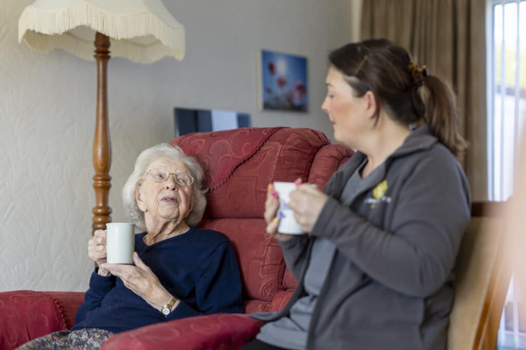 Elderly white lady in chair having a cup of tea with a carer from The EH Group